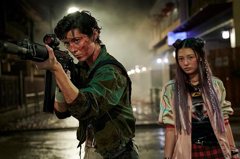 警戒から共闘へ、危険な夜をともに進む二人 映画『ケイト』で、銃を構えるケイトと、その後ろに立つ少女アニ